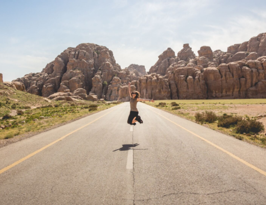 Young female adult jumping in the middle of a road with rocky mountains in the background